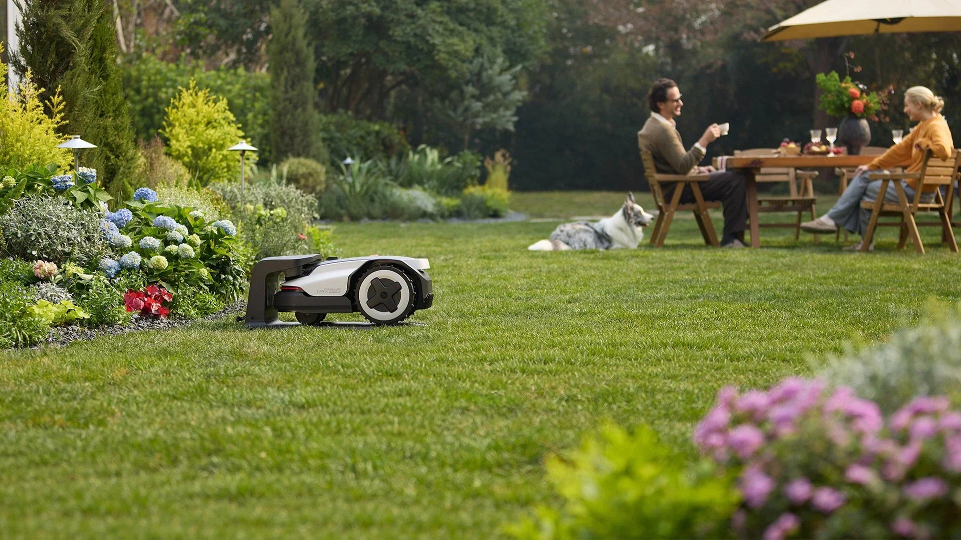 Wire-Free robot mower in it's charging station with family in the background on a freshly cut lawn.