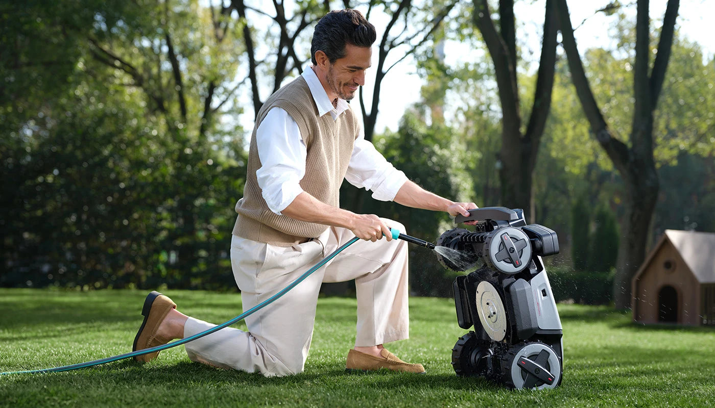 Person rinsing the underside and cutting deck of a RockMow robot mower with a garden hose.